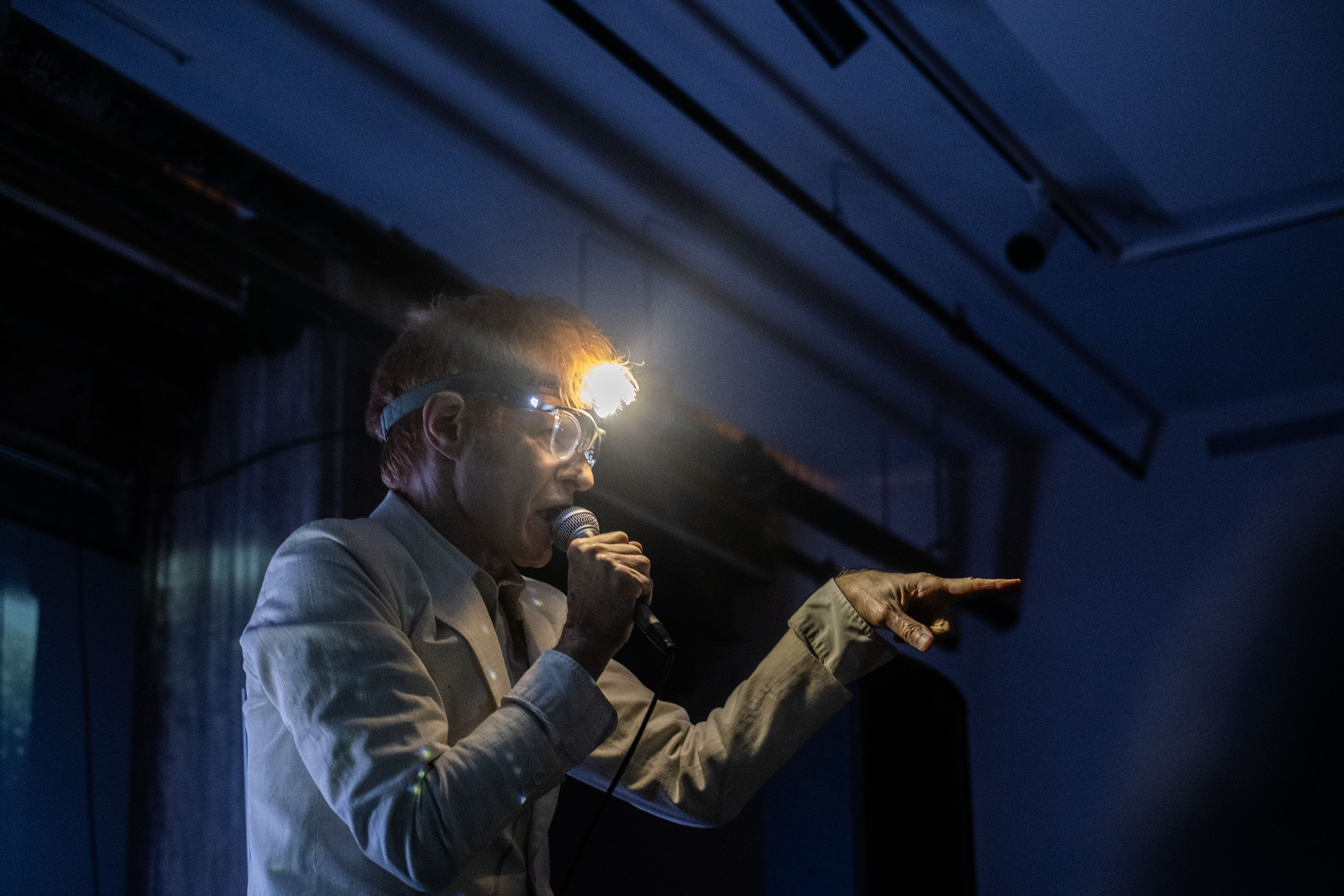 A performer, wearing a bright headlamp, sings into a microphone while pointing out towards the audience in a dimly lit room. 