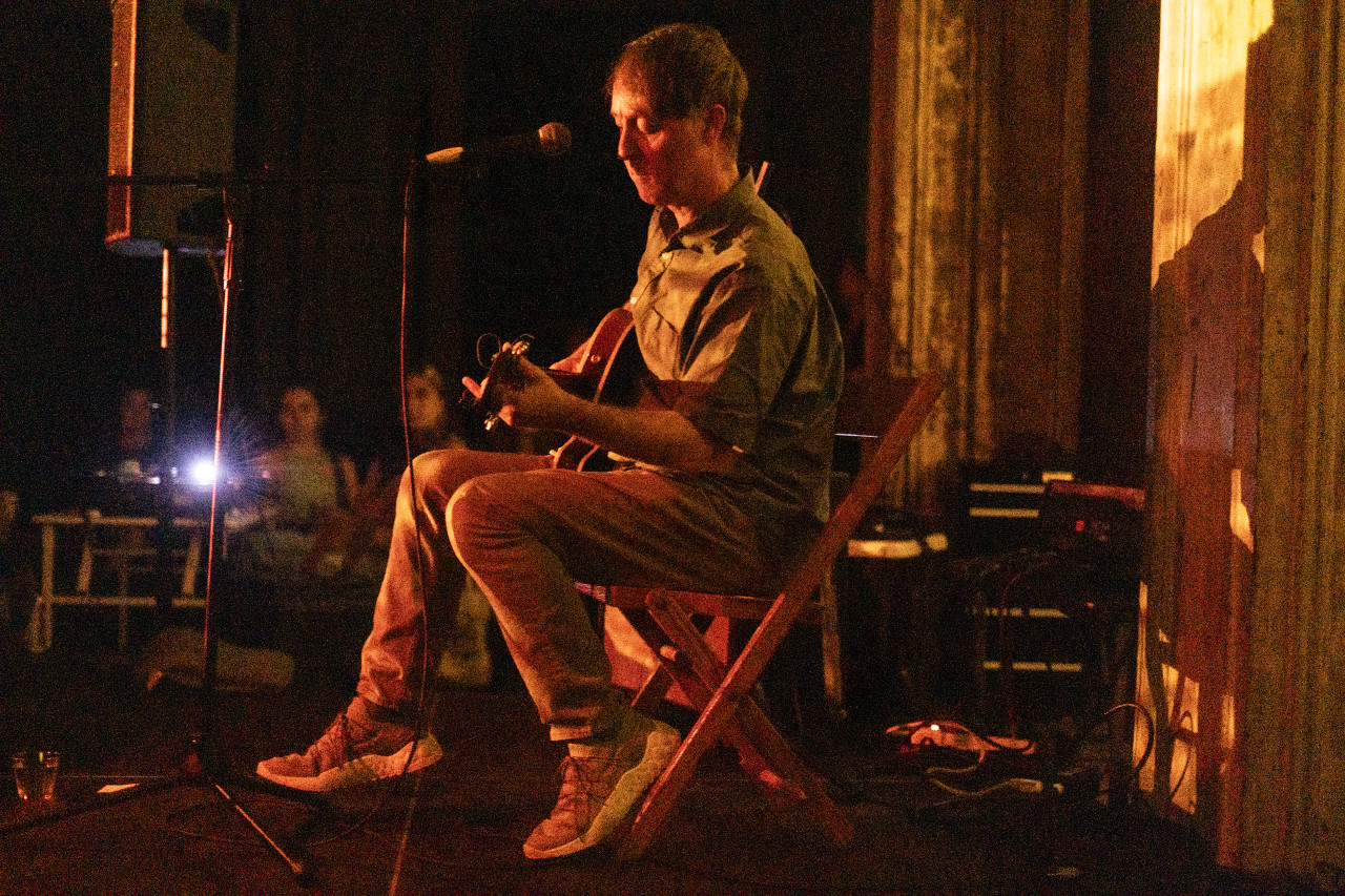 The left side of a performer is seen seated on stage, looking down as they play guitar. Warm yellow light shines upon them and a bright blue light flashes across the stage.