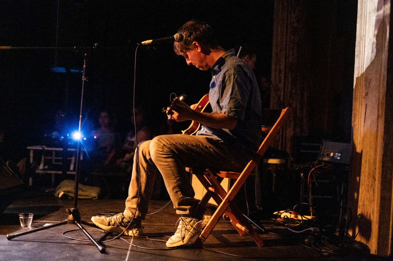 The left side of a performer is seen seated on stage, looking down as they play guitar. Warm yellow light shines upon them and a bright blue light flashes across the stage.