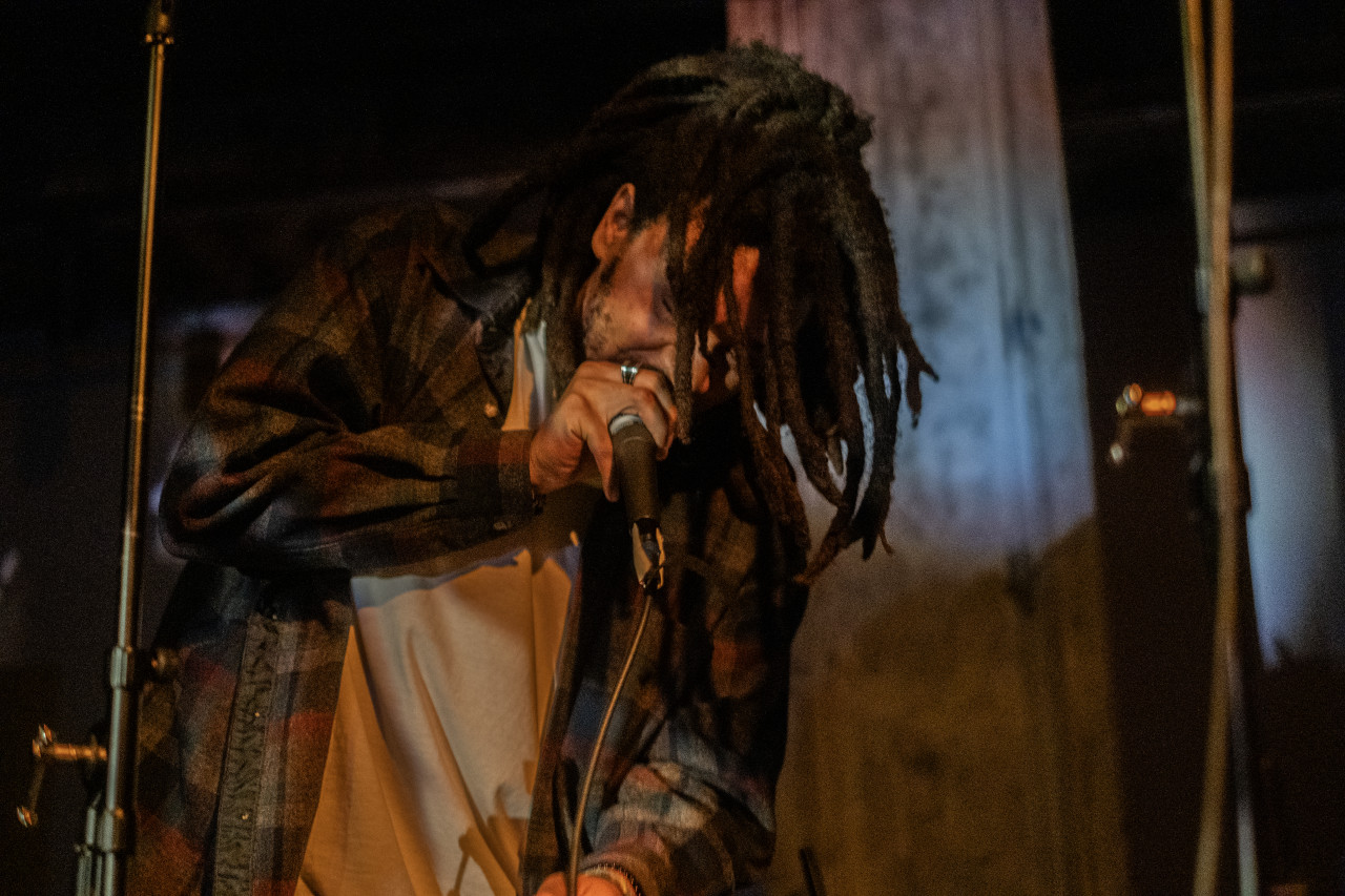 A performer grips the top part of a microphone close to their mouth. Their head is tilted downwards and hair falls over the front of their face. They are illuminated by a soft yellow light.