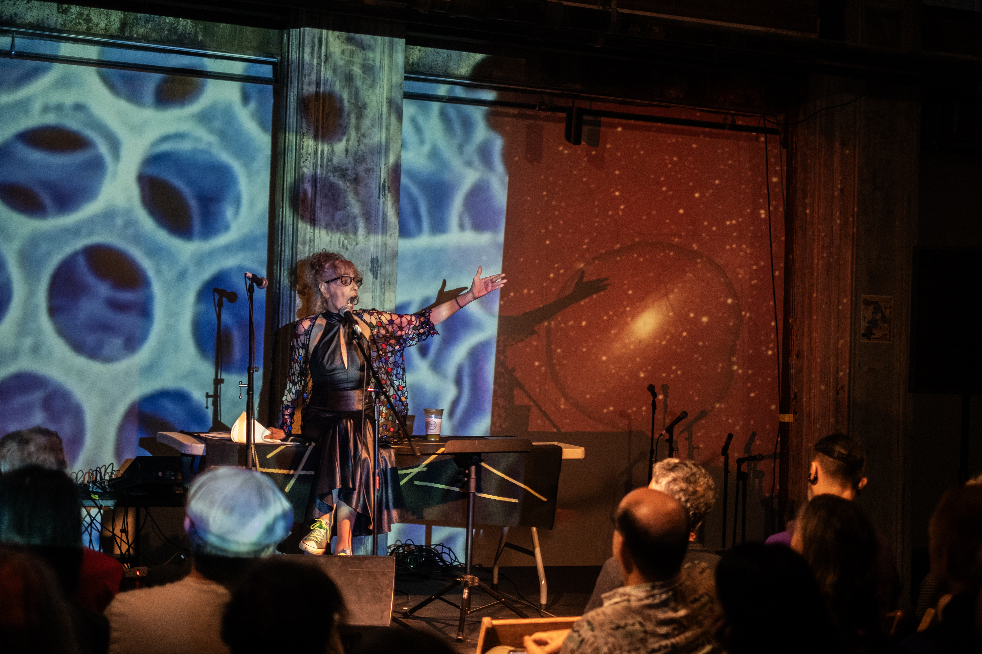 A seated performer in a black leather dress sings into a microphone, with their mouth open wide and left arm outstretched. On the wall behind, a projection of an abstract design of blue circles is juxtaposed by an orange-tinted photograph of a galaxy and stars.
