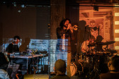 Three musicians perform on stage, lit by soft yellow light. The leftmost performer is seated at a table with mixing boards. The center performer plays trumpet into a microphone. The rightmost performer plays the drums. A light blue mottled projection shines on the left half of the wall and performers, while an orange projection of an illustrated skeleton shines on the right.