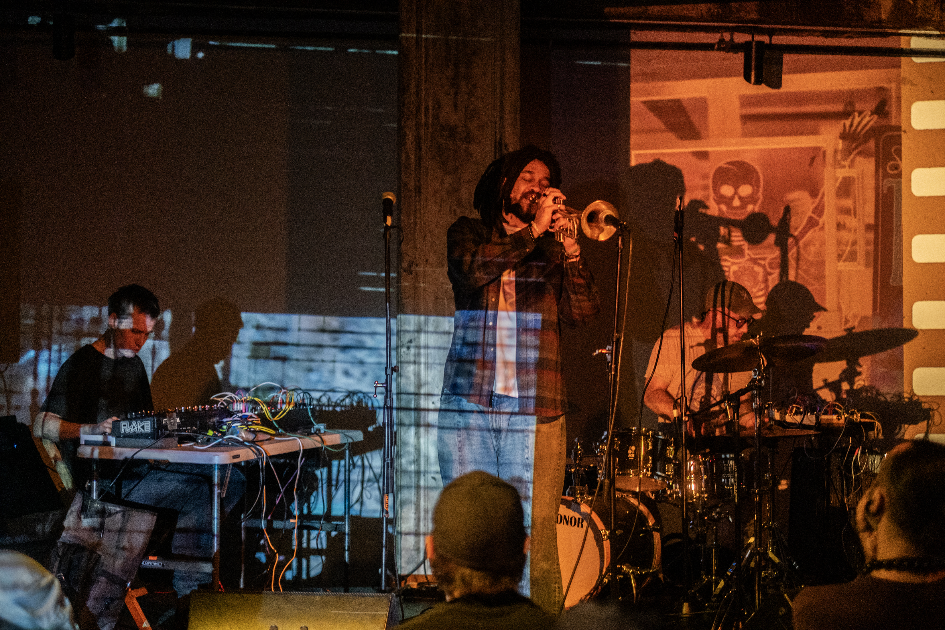 Three musicians perform on stage, lit by soft yellow light. The leftmost performer is seated at a table with mixing boards. The center performer plays trumpet into a microphone. The rightmost performer plays the drums. A light blue mottled projection shines on the left half of the wall and performers, while an orange projection of an illustrated skeleton shines on the right.