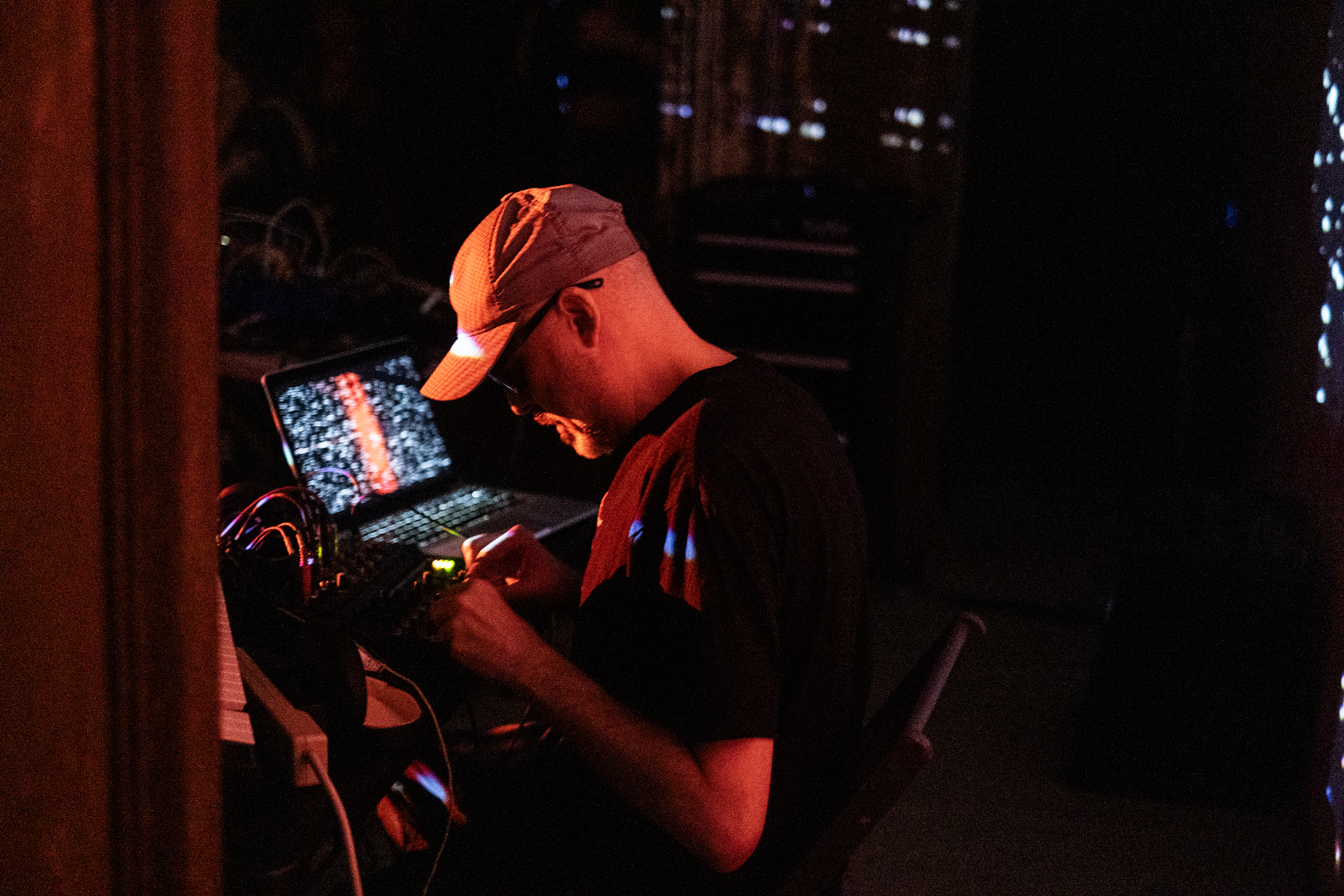 The left side of a performer wearing a baseball cap is bathed in red light as they adjust knobs on a mixing board. An illuminated laptop screen sits to their right.