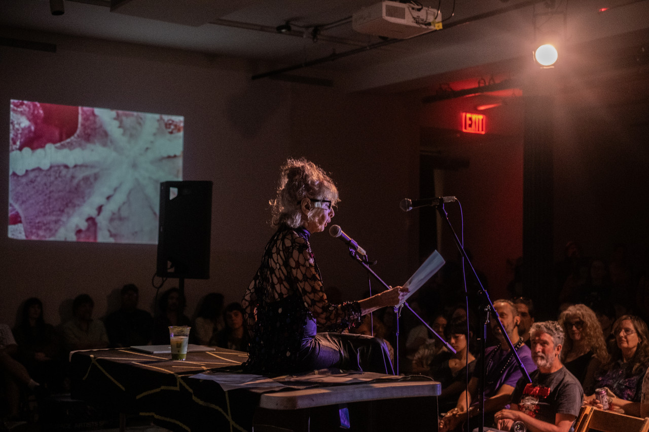 The right side of a performer is seen illuminated by a white light as they speak into a microphone. Some audience faces seated near the front are also visible. Behind a large speaker, a close-up image of a red, pink, and white starfish is projected onto the wall.