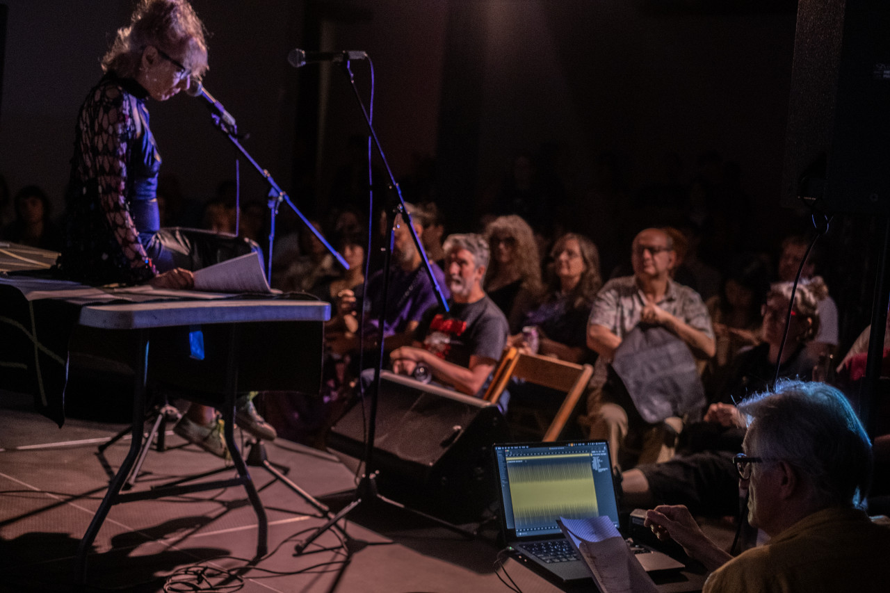 The right side of a performer, seated in front of a microphone, is seen as they look down at their notes. Audience faces are illuminated by the radiant stage light. In the right foreground, an individual watches a laptop screen.