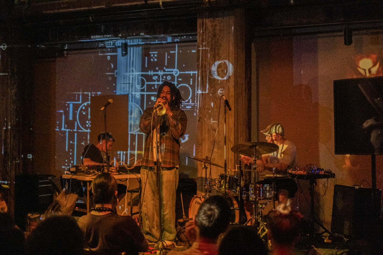 Three musicians perform on stage, lit by soft yellow light. The leftmost performer is seated at a table with mixing boards. The center performer plays trumpet into a microphone. The rightmost performer plays the drums. An abstract projection of blue circles, dots, and straight lines shines on the wall behind.