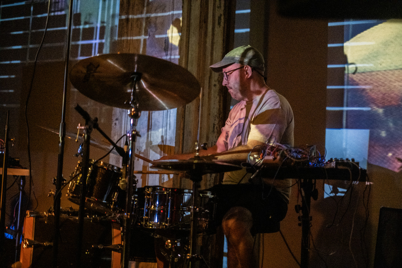 A performer wearing a baseball cap plays drums. A black, white, yellow, and green projection of horizontal and vertical lines shine on the performer and the wall behind.