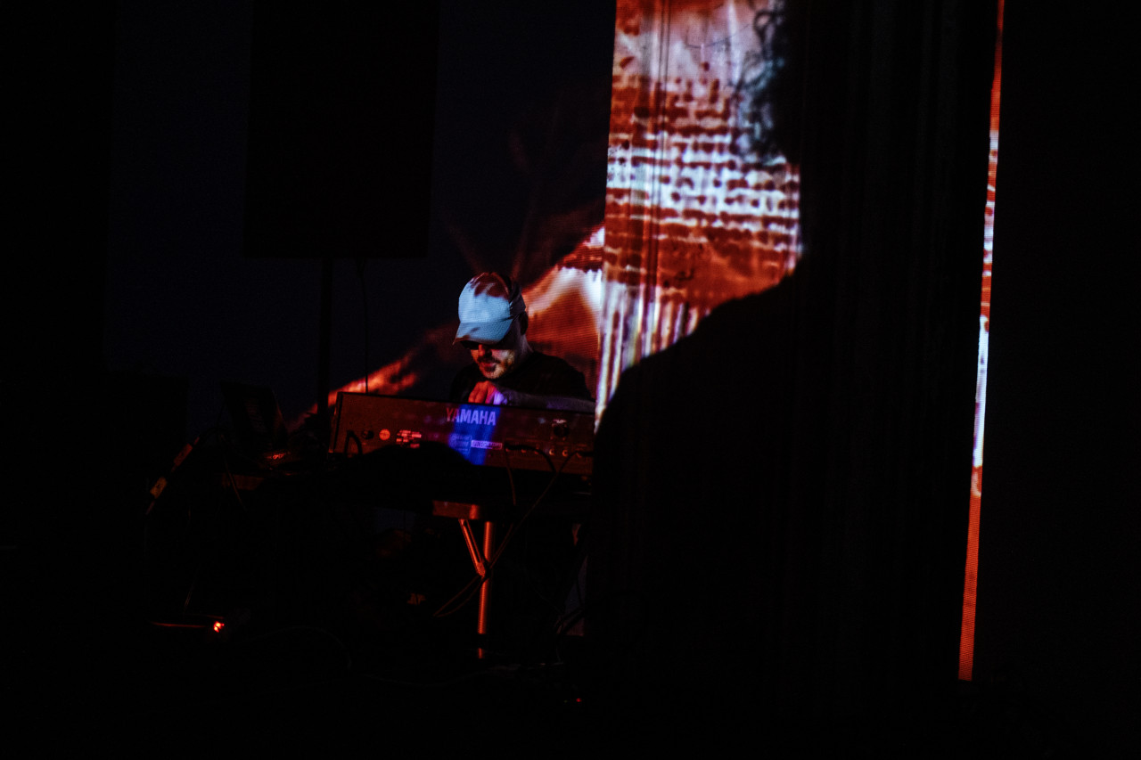 A performer wearing a baseball cap looks down at a Yamaha mixing board on a darkened stage. A red and white projection with an abstracted woven texture lights up their body and the wall behind.