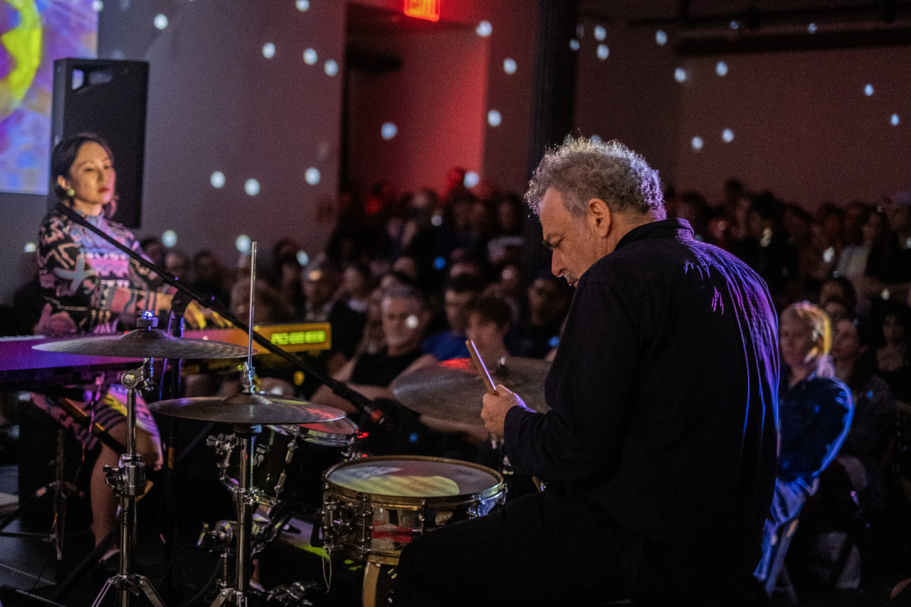 The backside of a seated performer playing drums is seen in focus on the right. On the left, out of focus, a seated performer plays the keyboard. In the background, faces of audience members are visible. The space is speckled with small dots of white light.