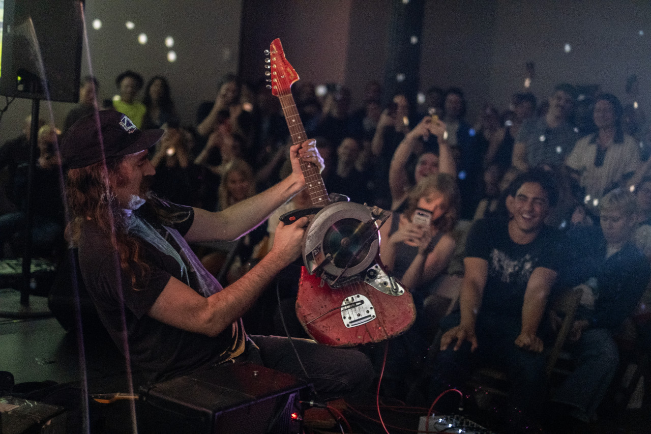 A seated performer grips the neck of a rugged bright red electric guitar in their left hand while holding a circular saw to the strings in their right hand. In the background, rows of seated audience members are seen smiling, and several hold their phones up to record. The space is speckled with small dots of white light.