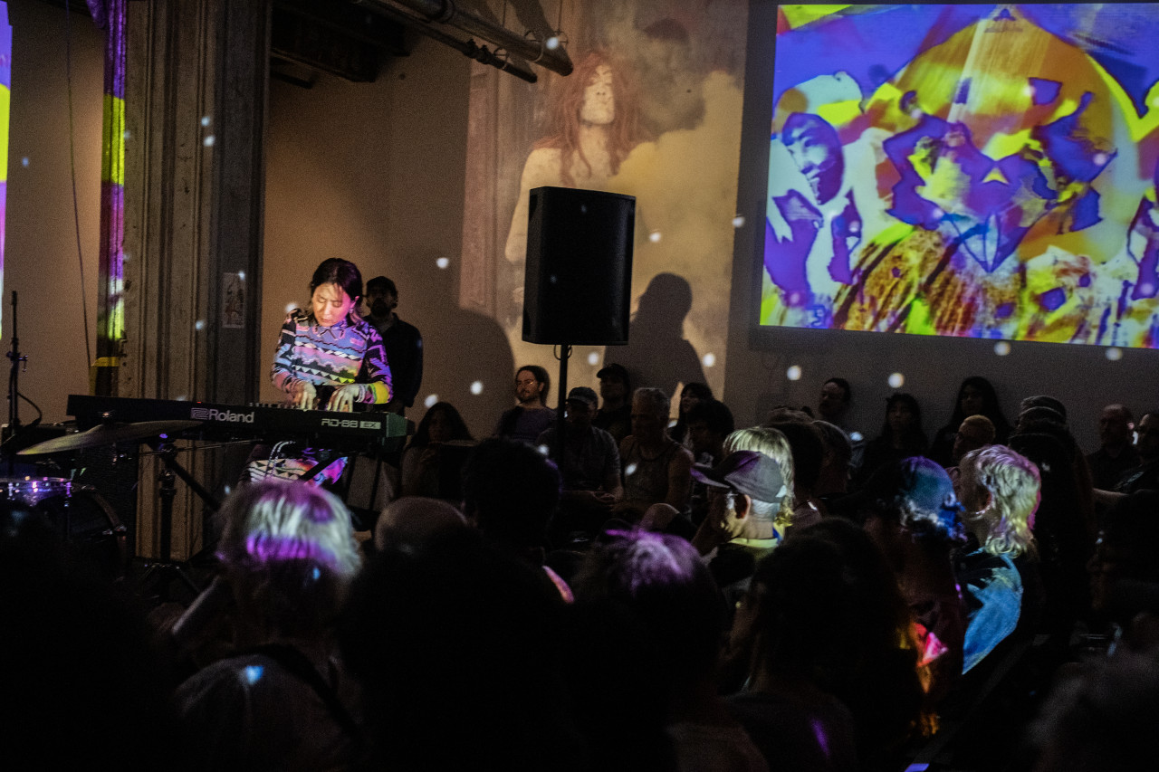 On the left, a seated performer looks down as they play the keyboard. On the right, rows of audience members are seated on chairs and along the right wall. On the left side of the wall behind, a projection of a figure with long orange hair surrounded by murky clouds is juxtaposed by an abstract purple and yellow design. White dots of light speckle the space around.