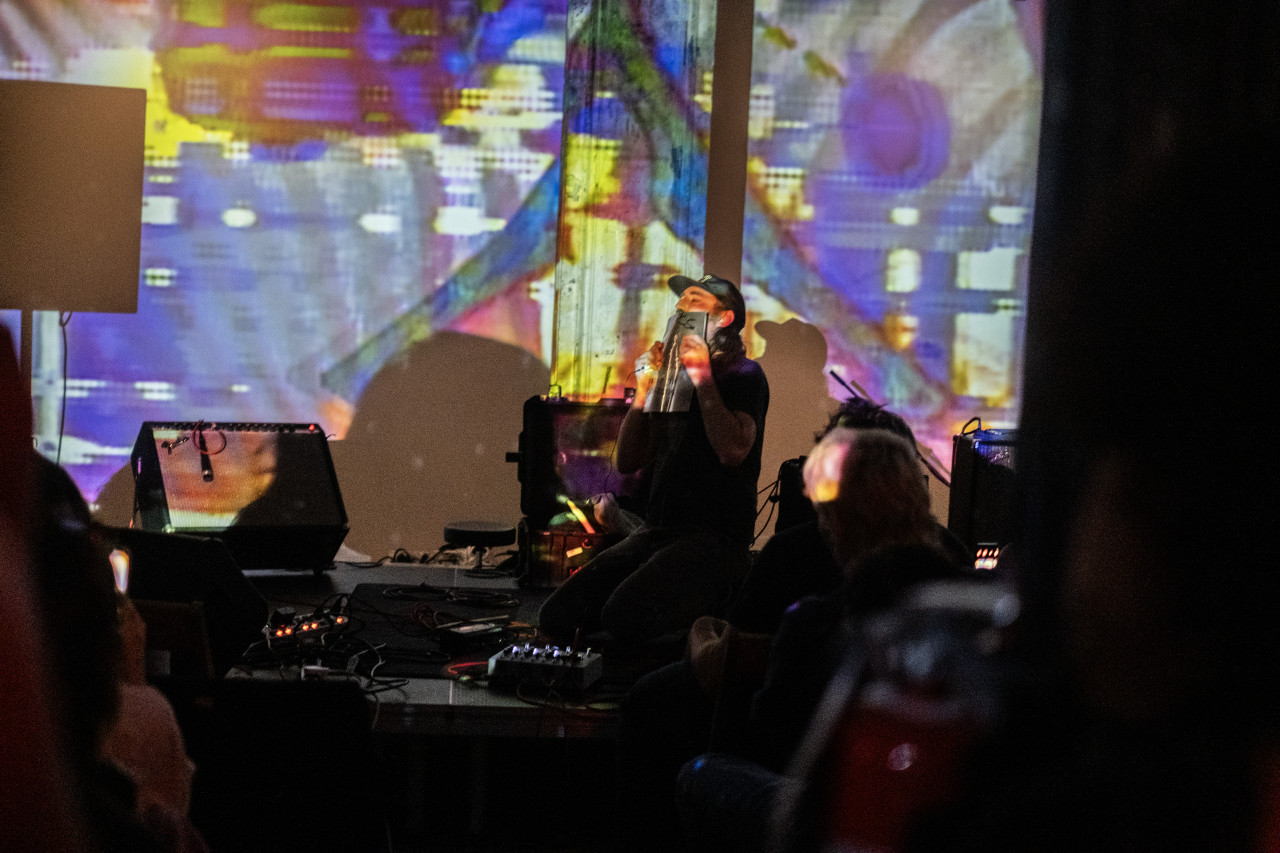 A performer kneels on stage with their eyes closed as they hold a thin metal sheet over their mouth. They are surrounded by amps, speakers, and mixing boards. An abstract yellow, blue, grey, and white projection shines on them and the wall behind.