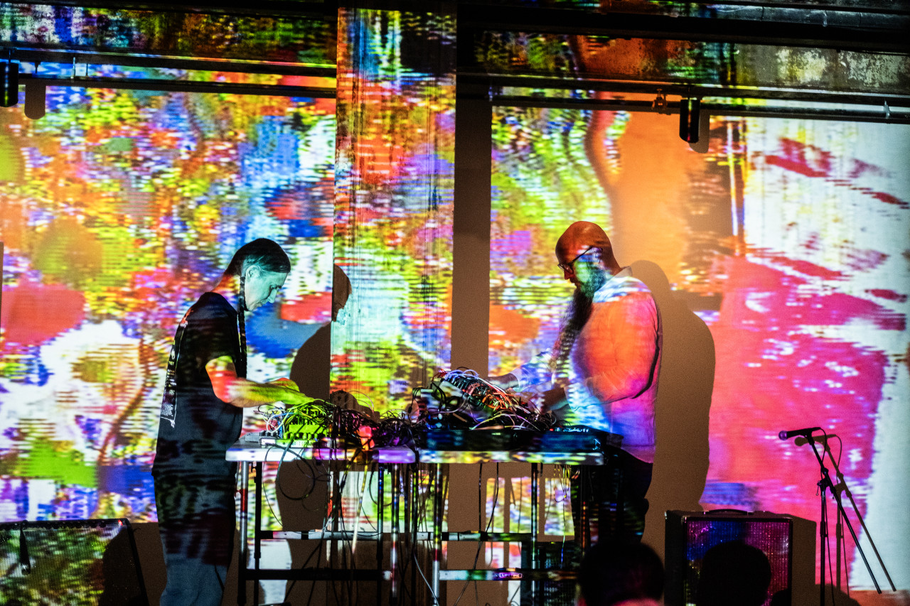 The sides of two performers are seen on stage as they stand at opposite ends of a table. They both gaze downwards where their hands adjust knobs on mixing boards. A yellow, red, orange, pink, green, purple, and white abstract pixelated projection shines on the performers and the wall behind.
