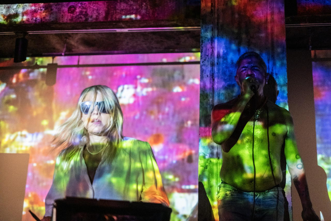 On the left, the top half of a performer is seen wearing sunglasses with their hair falling in front of their face. On the right, a shirtless performer is seen from the waist-up as they stand in front of a column with their eyes closed and microphone to their mouth. A blue, yellow, pink, purple, and green cloud-like pattern is projected onto the performers and the wall behind.