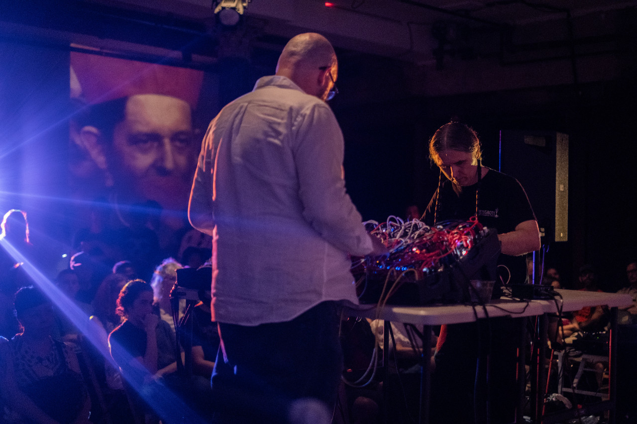 Two performers stand at opposite ends of a table as they gaze downwards to adjust knobs on mixing boards. One performer