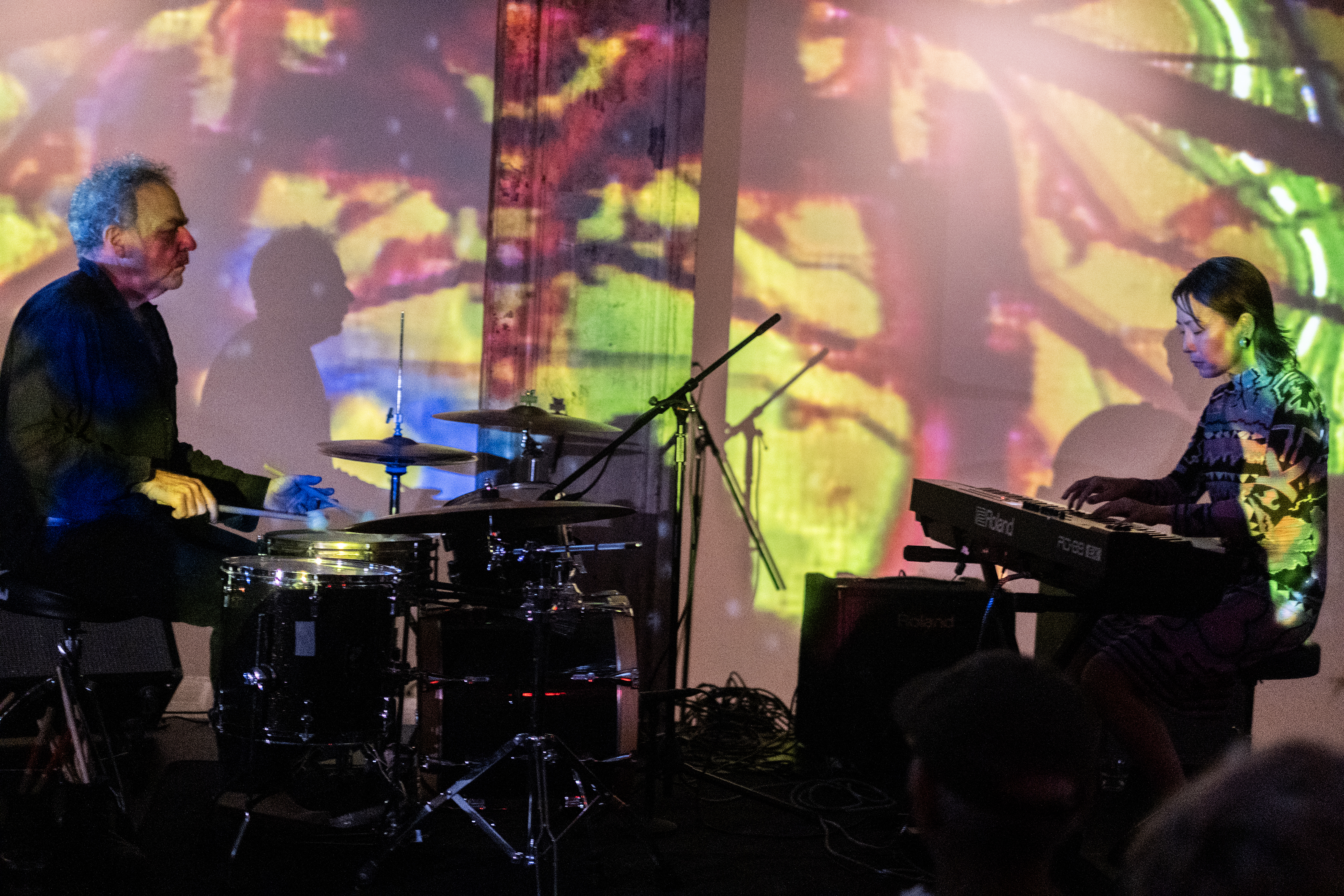 Two seated performers face each other on stage. The left performer plays the drums and the right performer plays a keyboard. An abstract yellow, red, and green projection shines on them and the wall behind.