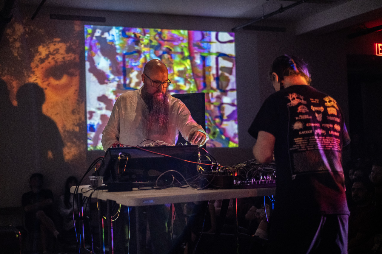 Two performers stand at opposite ends of a table as they gaze downwards to adjust knobs on mixing boards. One performer