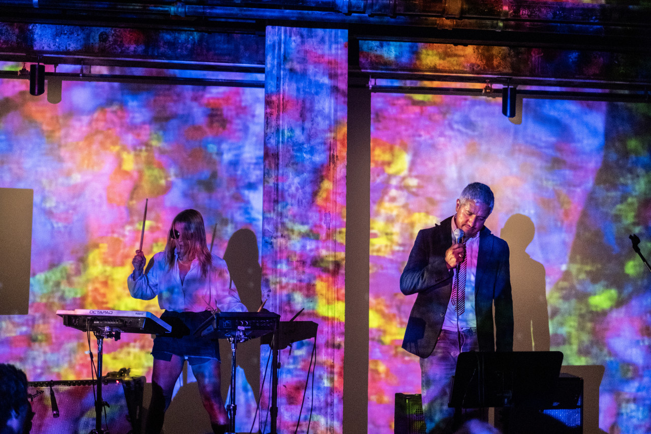 A performer on the left wears sunglasses as they play a drum pad, and a performer on the right holds a microphone to their mouth as they look down to a music stand. A blue, yellow, pink, purple, and green cloud-like pattern is projected onto the performers and the wall behind.