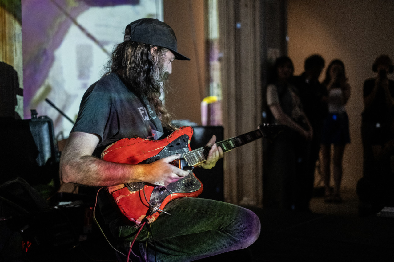 A seated performer rests a rugged bright red electric guitar on their leg while holding a walkie talkie in their right hand. Their face is turned to the left as they look down to the neck of the guitar. A purple and white projection shines on the performer and the wall behind. Several audience members stand in the background watching.