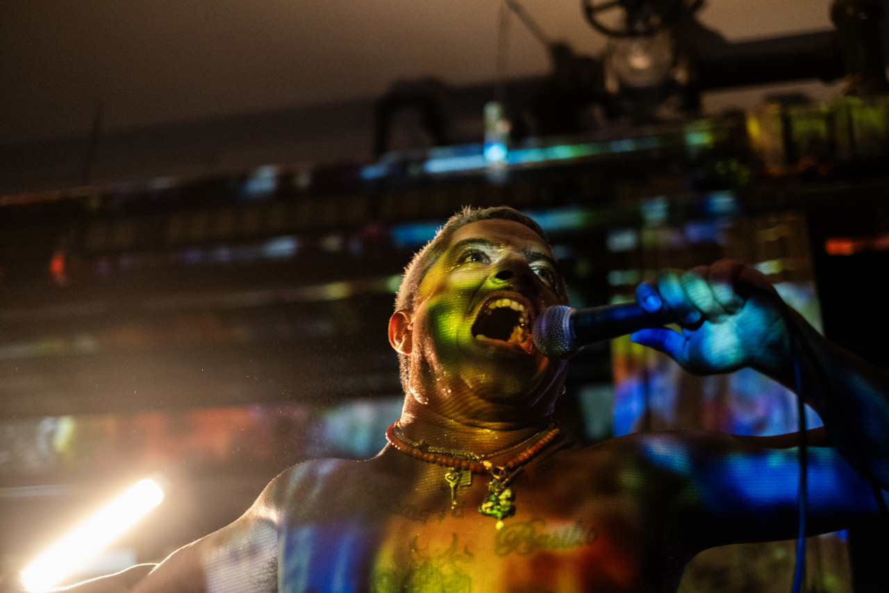 The face of a shirtless performer holding a microphone to their mouth is seen from a low angle. Their mouth is open wide as they look outwards. A green, yellow, and blue projection shines on them and the wall behind. A bright bar of yellow light shines on the left.