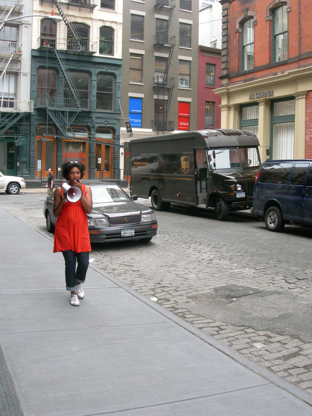 A person walking down a city street, speaking into a megaphone. Behind her are several cars and a truck, and she is surrounded by loft buildings.