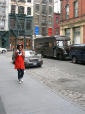 A person walking down a city street, speaking into a megaphone. Behind her are several cars and a truck, and she is surrounded by loft buildings.