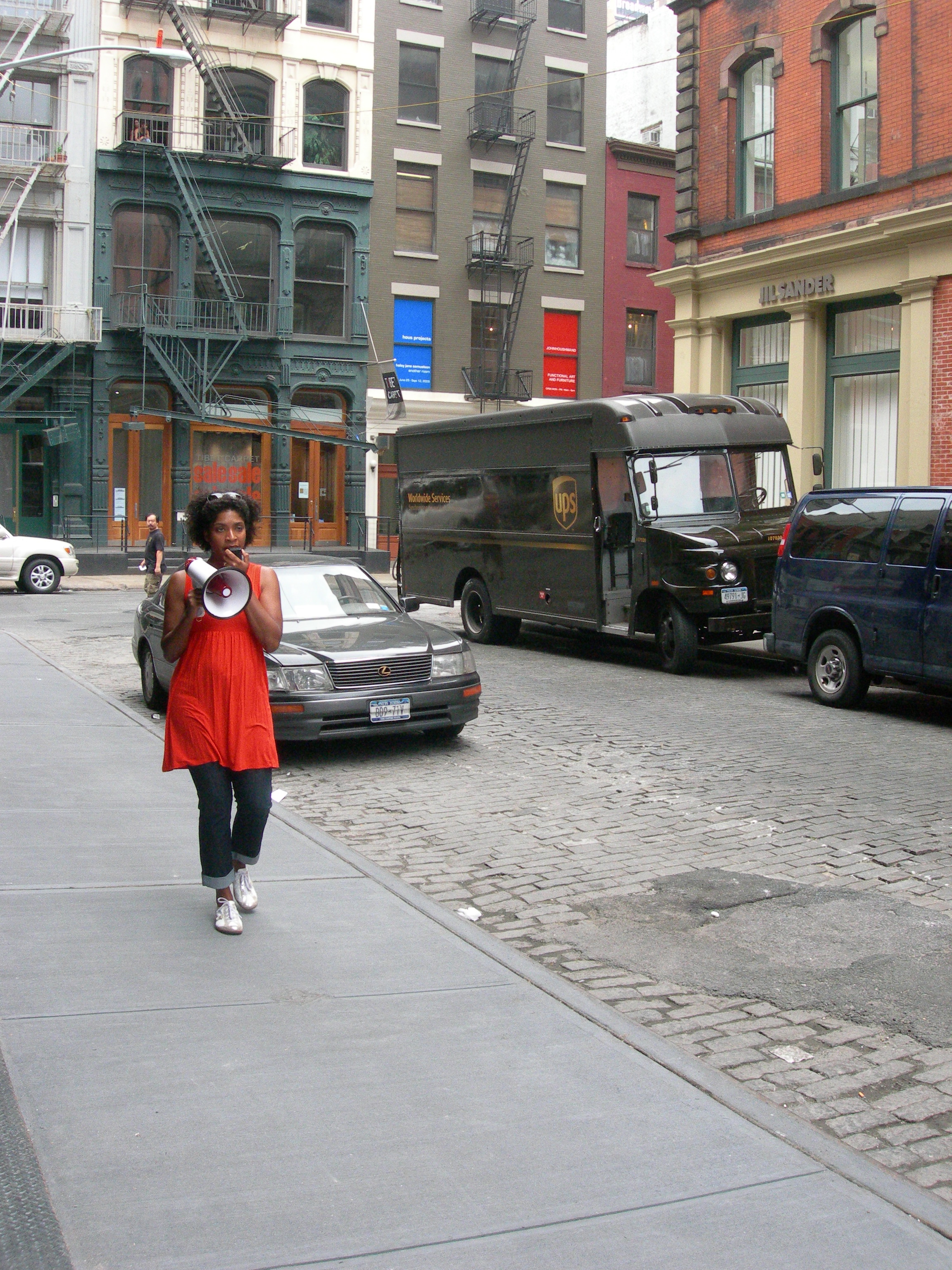 A person walking down a city street, speaking into a megaphone. Behind her are several cars and a truck, and she is surrounded by loft buildings.