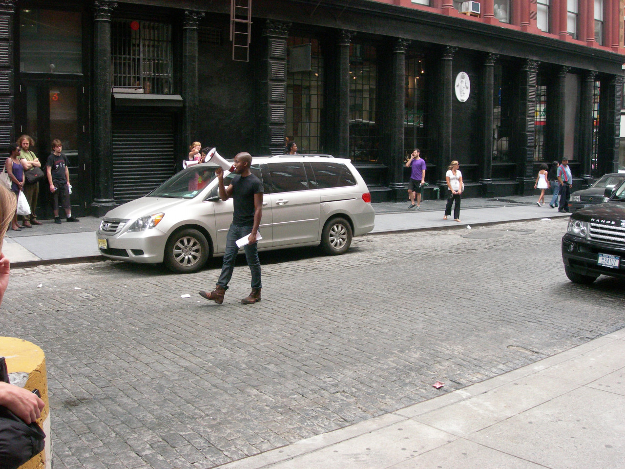 A person walking down a city street, speaking into a megaphone. Around him are several cars and scattered onlookers.