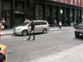 A person walking down a city street, speaking into a megaphone. Around him are several cars and scattered onlookers.