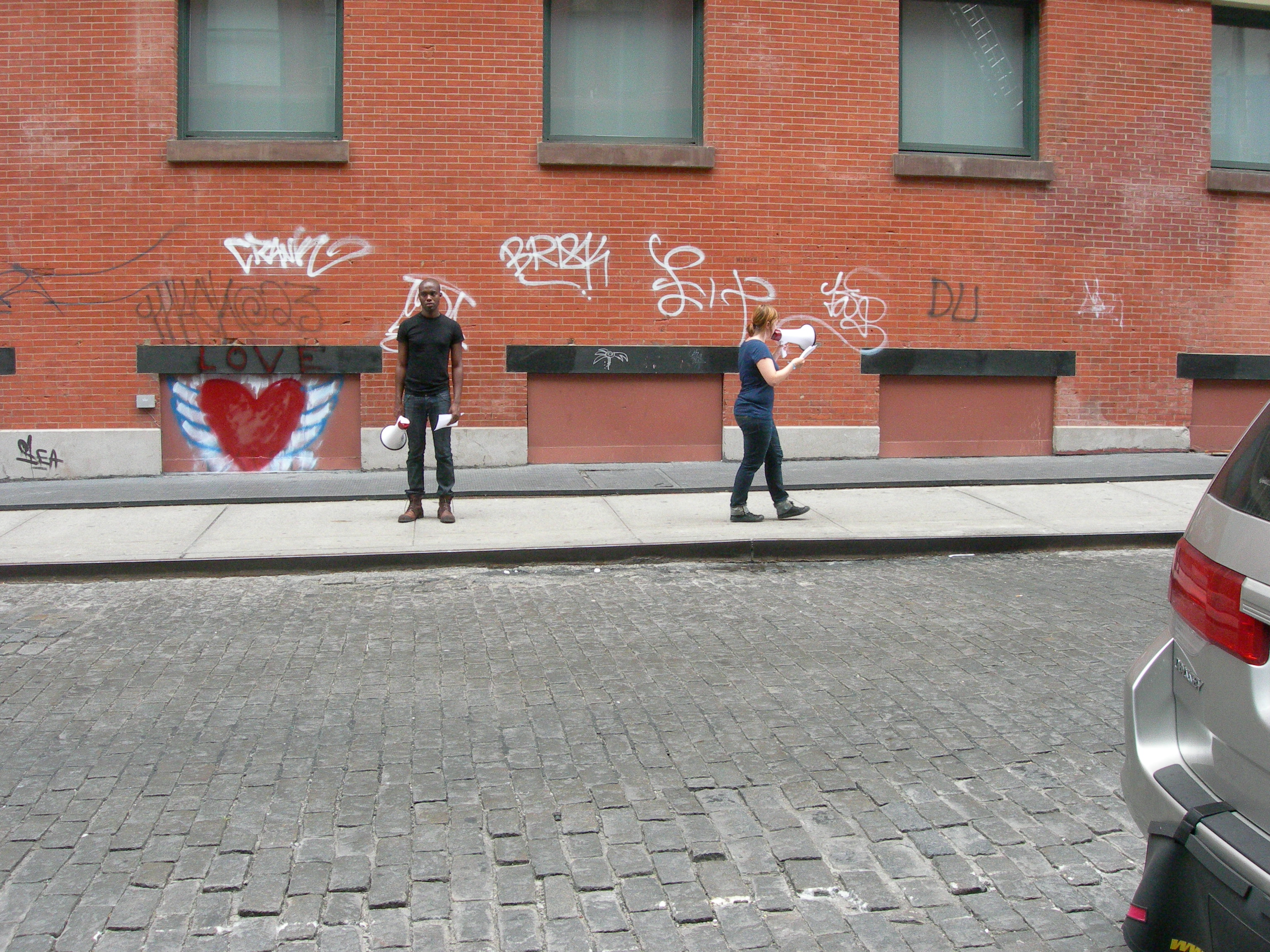 Two people on a city street in front of a graffitied brick wall. One faces the camera, holding a megaphone and a piece of paper at his sides. The other is walking, facing the side, and speaking into her megaphone and reading off a sheet of paper.