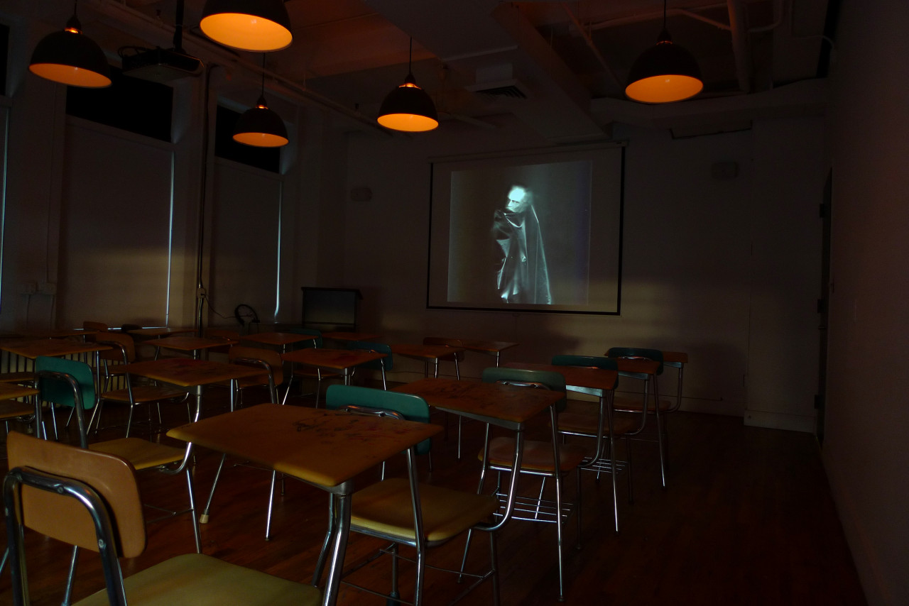 A darkened classroom, lit dimly with overhead orange lights. Three rows of chairs and desks face an image projected on a screen. The image is a blurred figure with a pale face enveloped in a black cape.