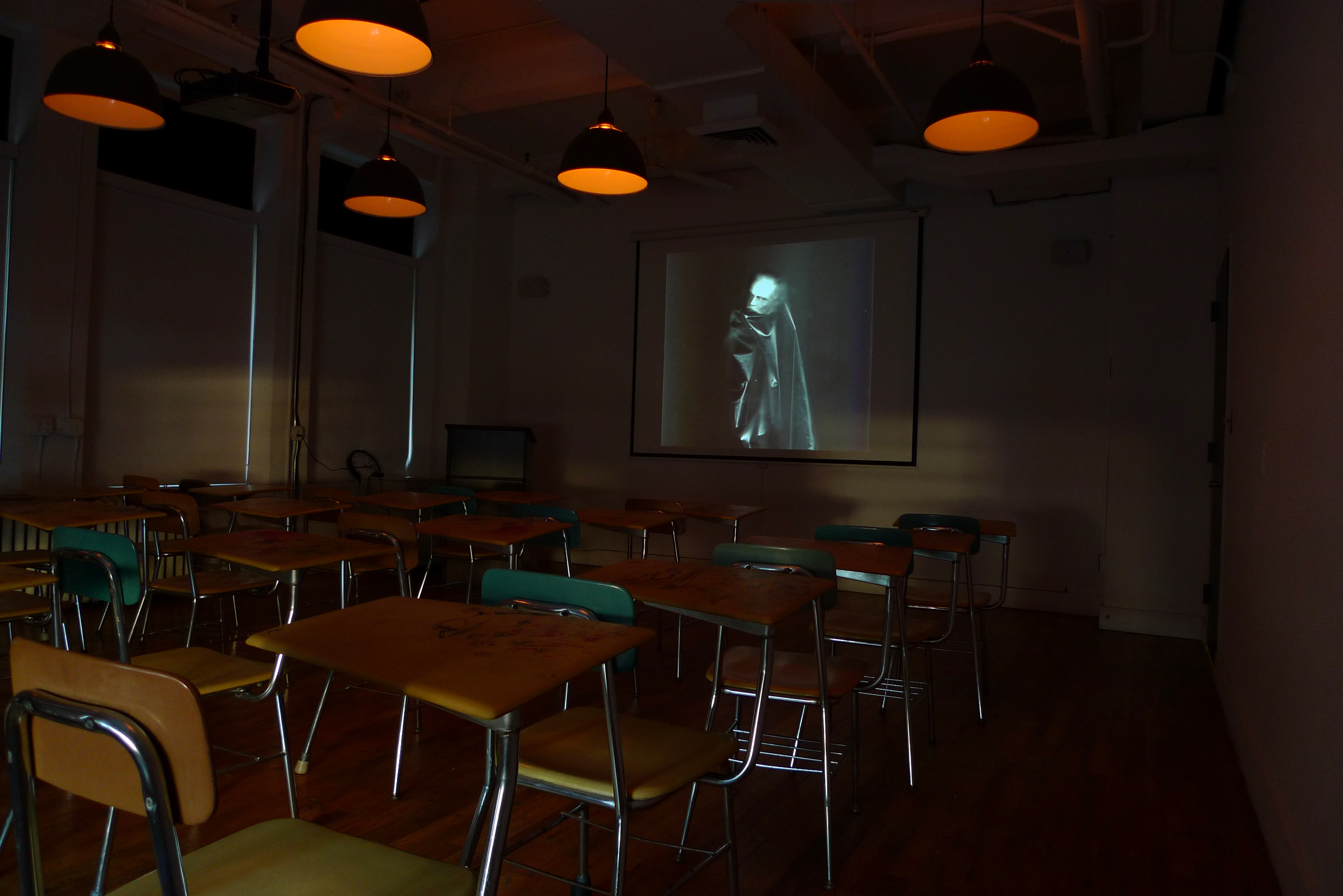 A darkened classroom, lit dimly with overhead orange lights. Three rows of chairs and desks face an image projected on a screen. The image is a blurred figure with a pale face enveloped in a black cape.