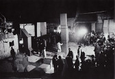 Black and white image of a crowd of people gathered in a large open space. Several hollow, metal sculptures composed of geometric components are installed in the space. Some performers interact with the sculptures—one figure walks across one of the metal forms that lays on the ground.