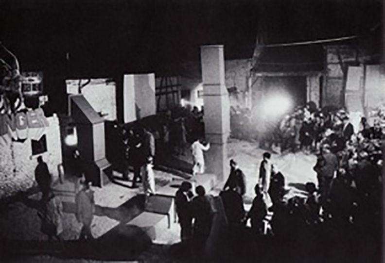 Black and white image of a crowd of people gathered in a large open space. Several hollow, metal sculptures composed of geometric components are installed in the space. Some performers interact with the sculptures—one figure walks across one of the metal forms that lays on the ground.