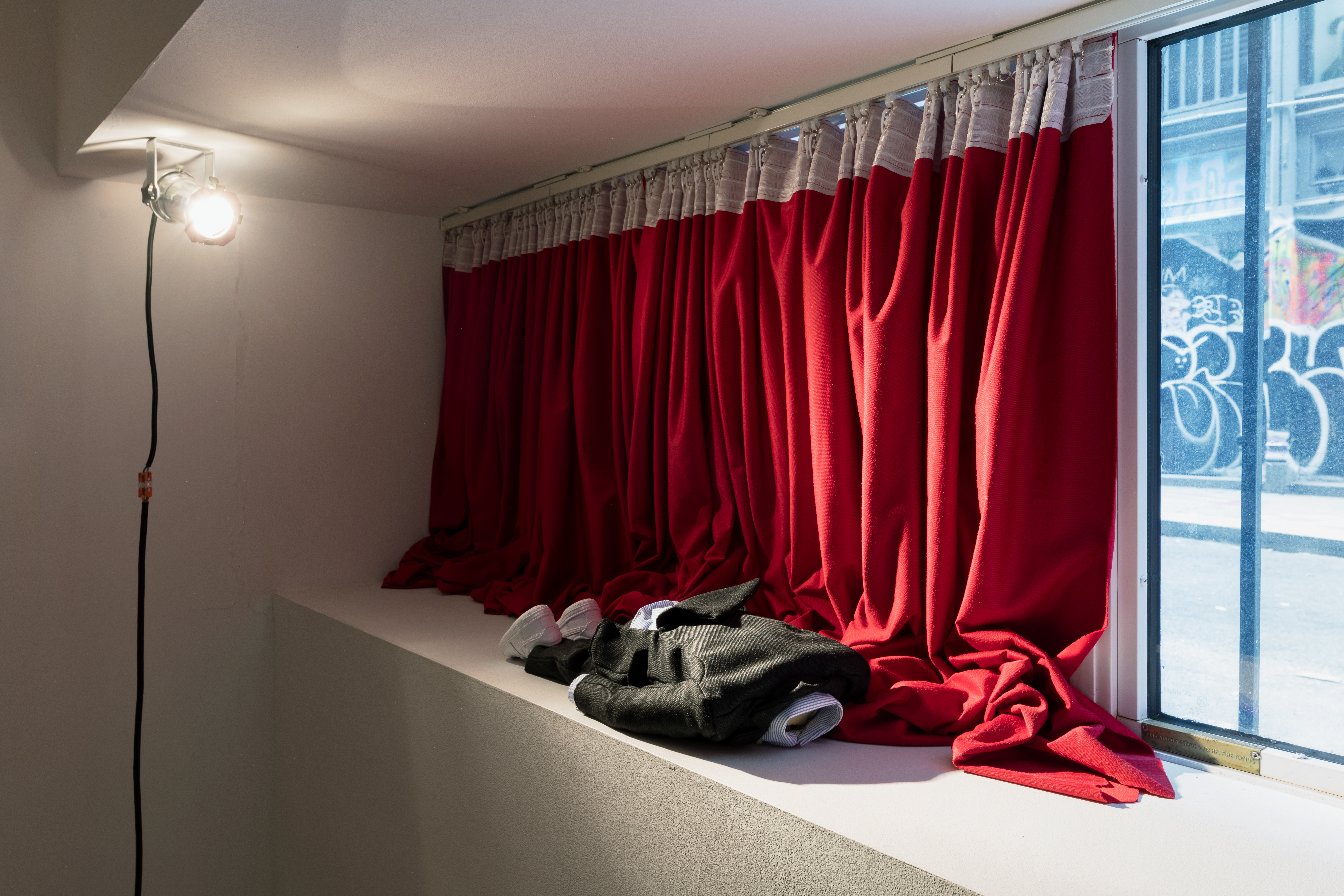 A marionette wearing a black suit, collared shirt, and white Nike Air Force 1s is positioned laying prone on a windowsill. Red curtains cover half of the window behind the marionette. In the corner, a small stage lights is directed at the marionette. Outside of the window is an alleyway with graffiti.