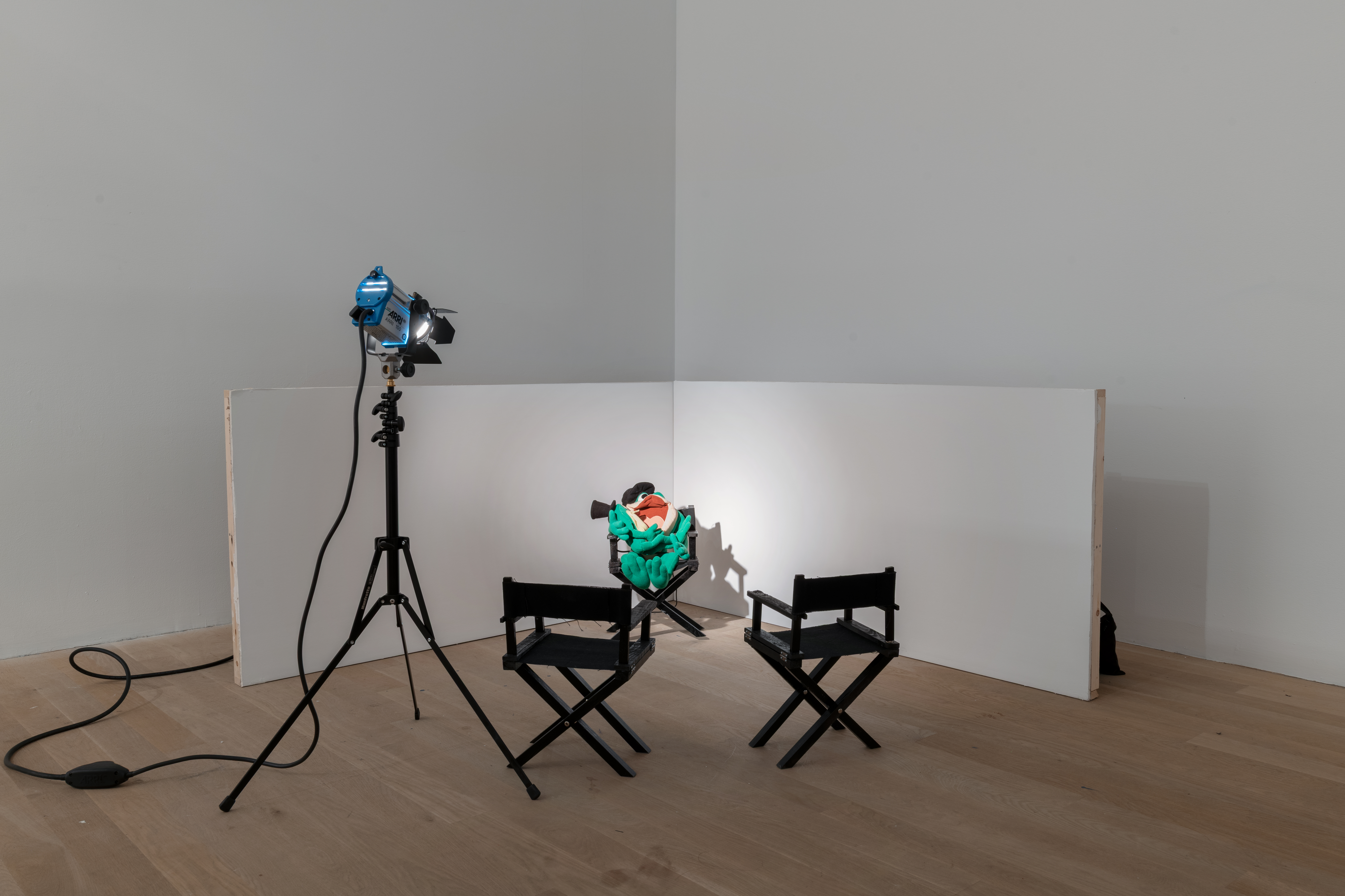 Two pieces of wood are joined to create a miniature stage set. In the center, sitting in a miniature director's chair are two cartoon frog plushies wearing black top hats. The larger frog plushy cradles the smaller frog in it's arms. Two more director's chairs are positioned facing the plushies, along with a miniature stage light and stand that shines a spotlight on the plushies.