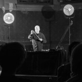 Black-and-white photograph of a figure performing on stage. The figure is standing, manipulating the bow of a string instrument. Layed out on the table in front of him is a laptop and various audio devices.