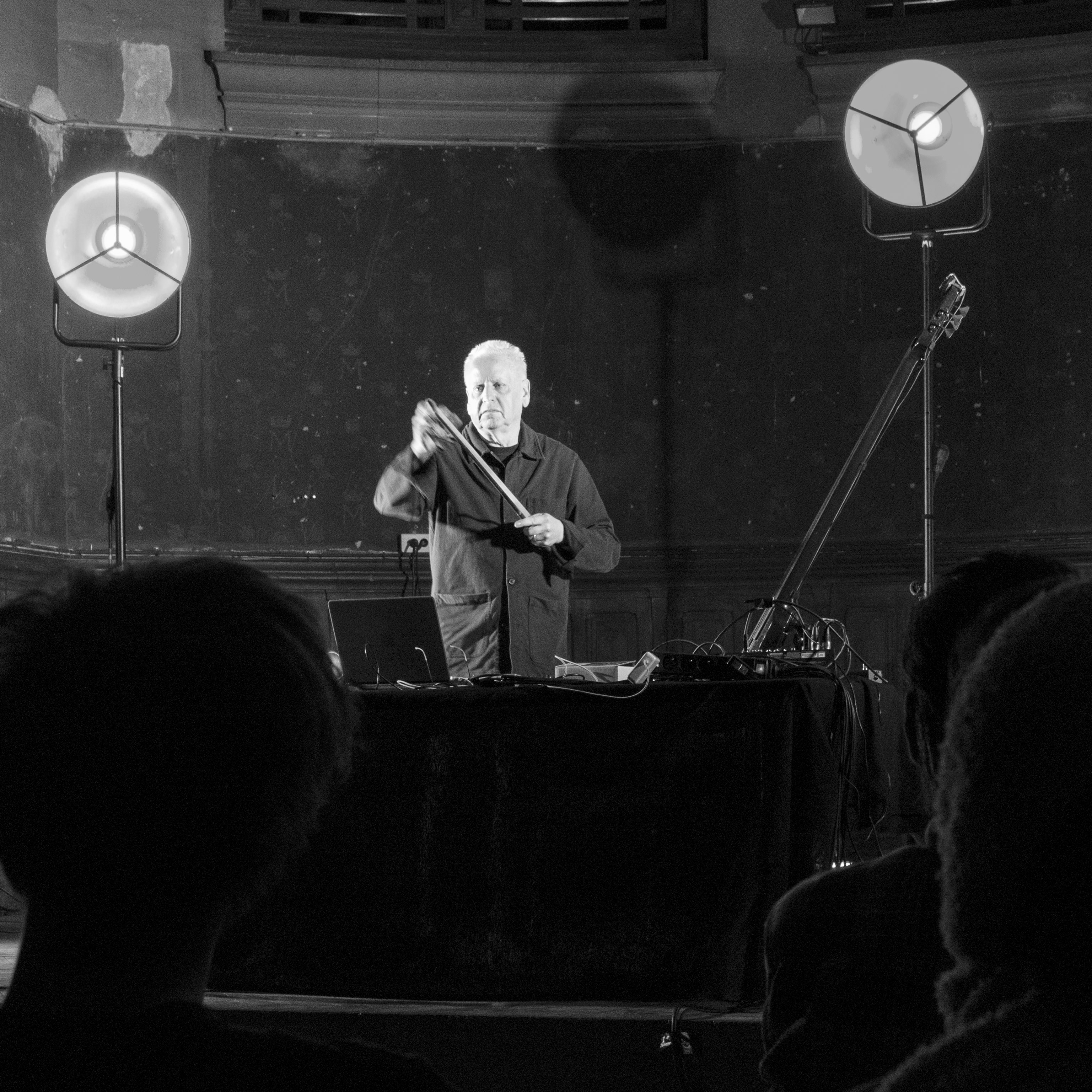 Black-and-white photograph of a figure performing on stage. The figure is standing, manipulating the bow of a string instrument. Layed out on the table in front of him is a laptop and various audio devices.