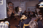 A crowd sits in a semi-circle arrangement of wooden chairs within a gallery space. In front of the audience, a man sits at a white table and reads from a piece of paper while speaking into a microphone. Directly behind the man is a large statue of a baby with glowing blue eyes.