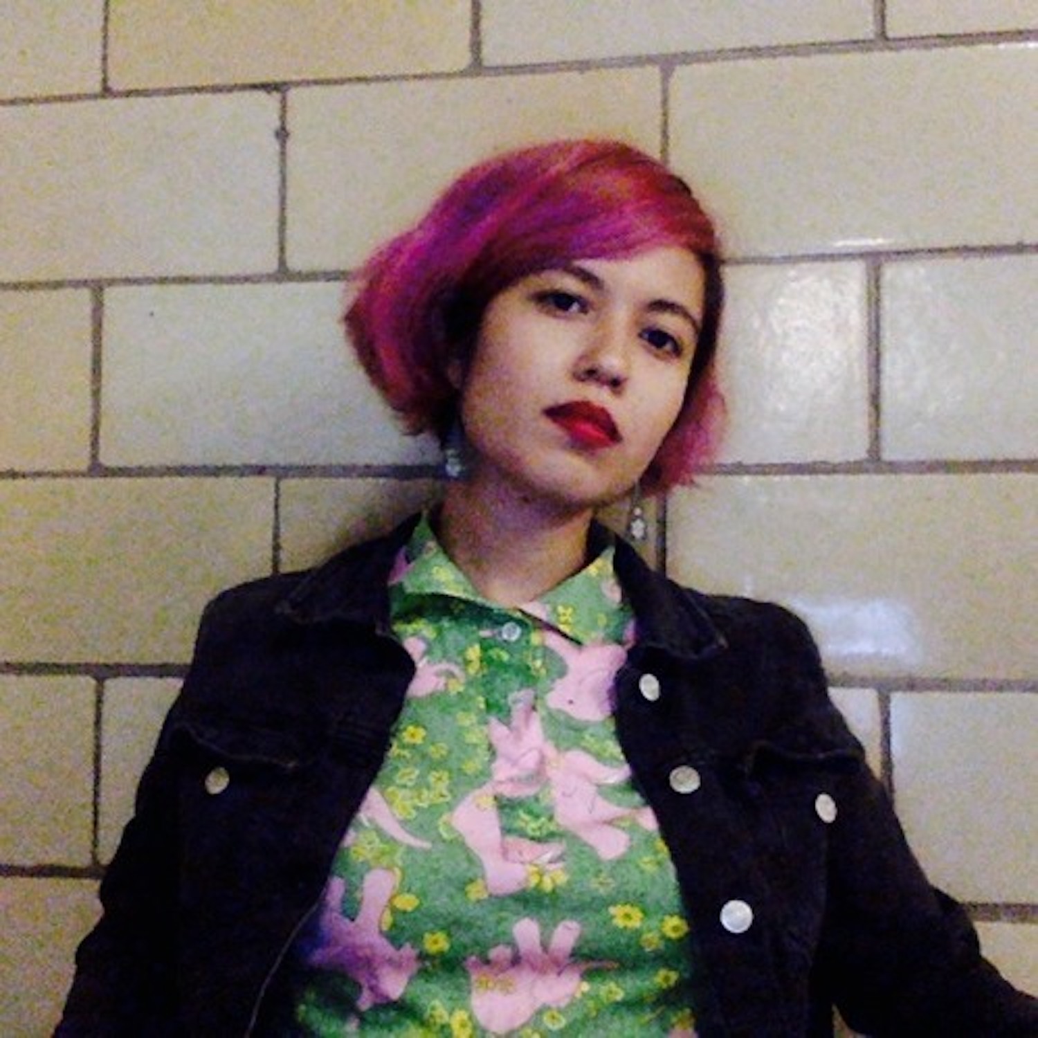 A figure, centered within the frame, leans against a beige tiled wall and stares directly into the camera.