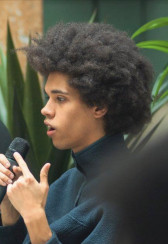 A figure, sitting in profile and centered within the frame, speaks into a microphone.