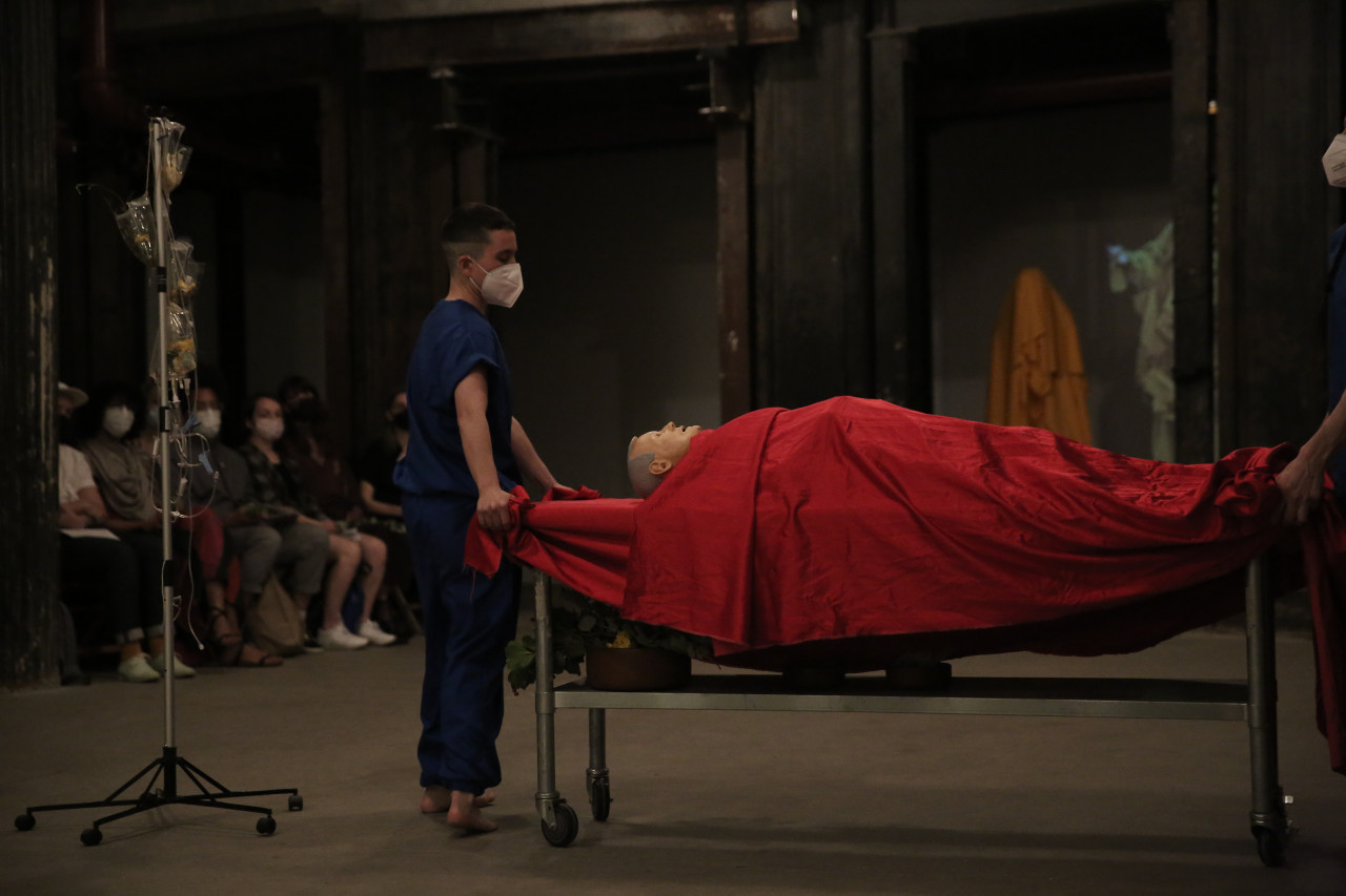 A person dressed in blue stands next to a mannequin draped in red fabric lying on a rolling table. To the left is an IV stand and people seated in the background.