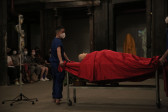 A person dressed in blue stands next to a mannequin draped in red fabric lying on a rolling table. To the left is an IV stand and people seated in the background.