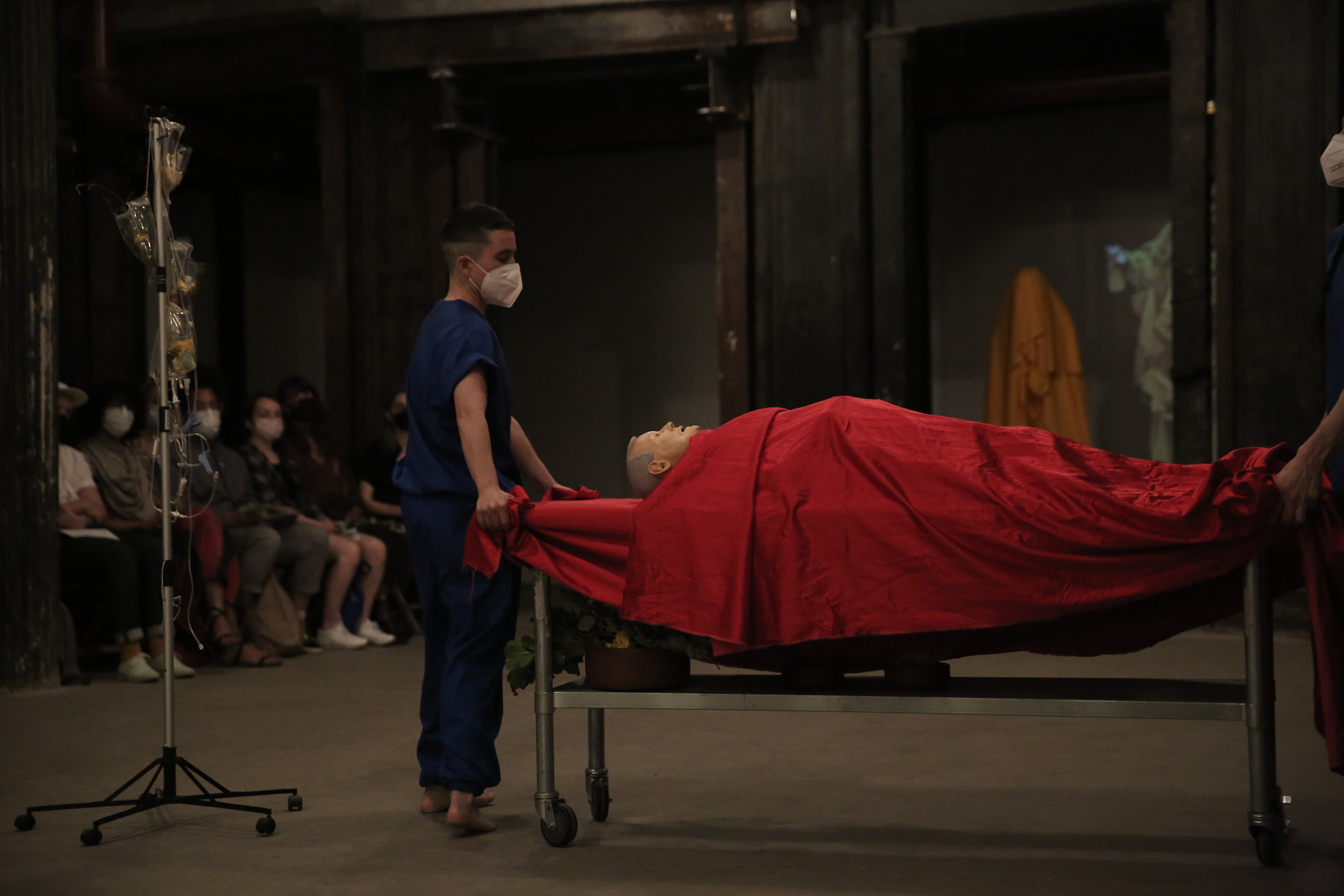 A person dressed in blue stands next to a mannequin draped in red fabric lying on a rolling table. To the left is an IV stand and people seated in the background.