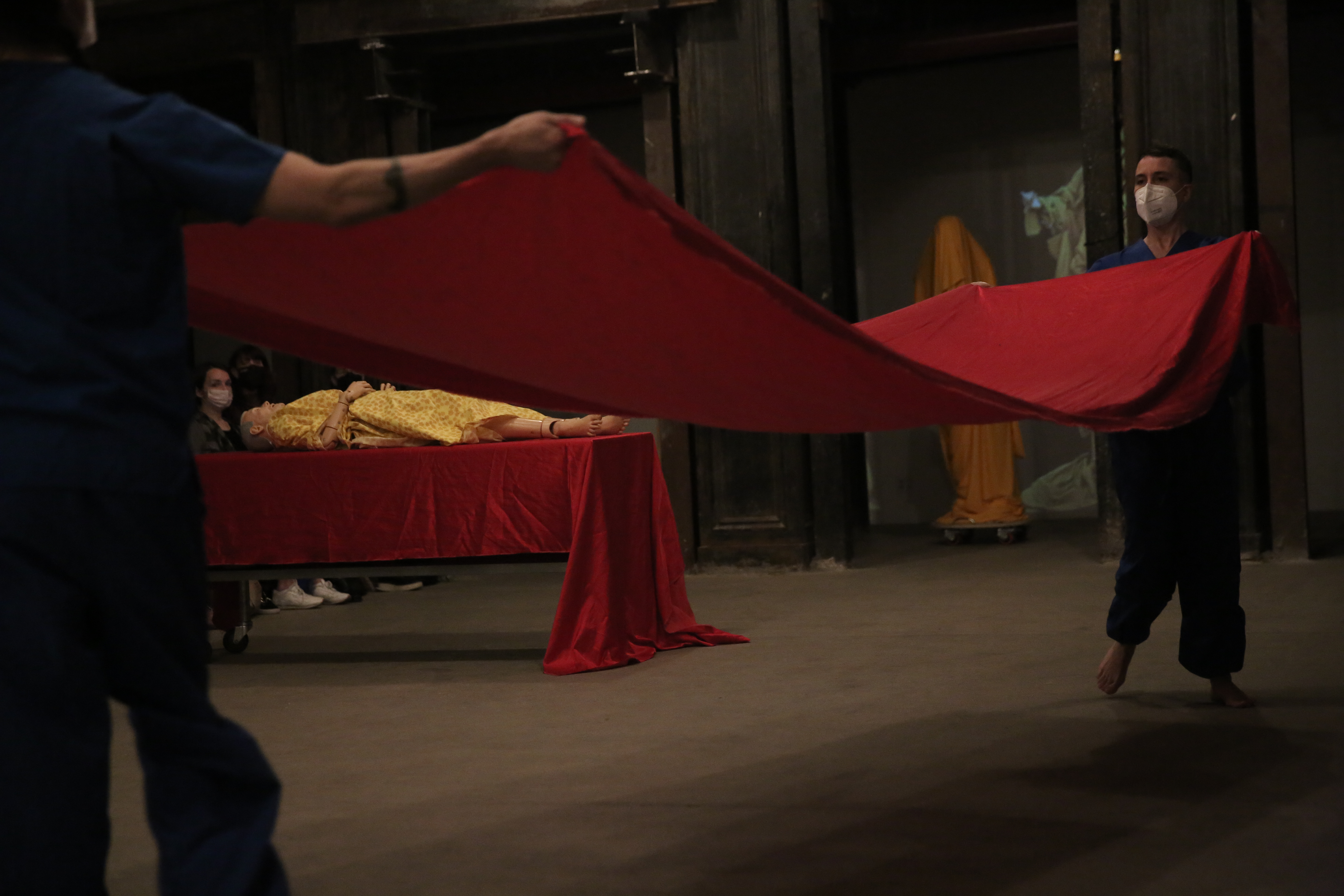 Two figures dressed in blue are holding a red fabric between them. In the distance, a manneqin in yellow clothing is lying on a table covered in red fabric. To the right, orange and yellow draped figures appear in the background.