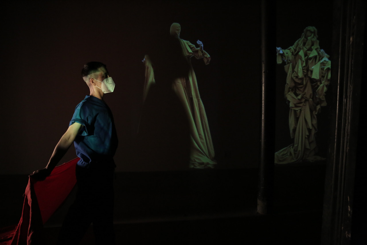 In a darkened space, a person dressed in blue faces to the right holding red fabric behind them. On the back wall are images of figures draped in yellow fabric.