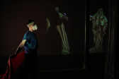 In a darkened space, a person dressed in blue faces to the right holding red fabric behind them. On the back wall are images of figures draped in yellow fabric.