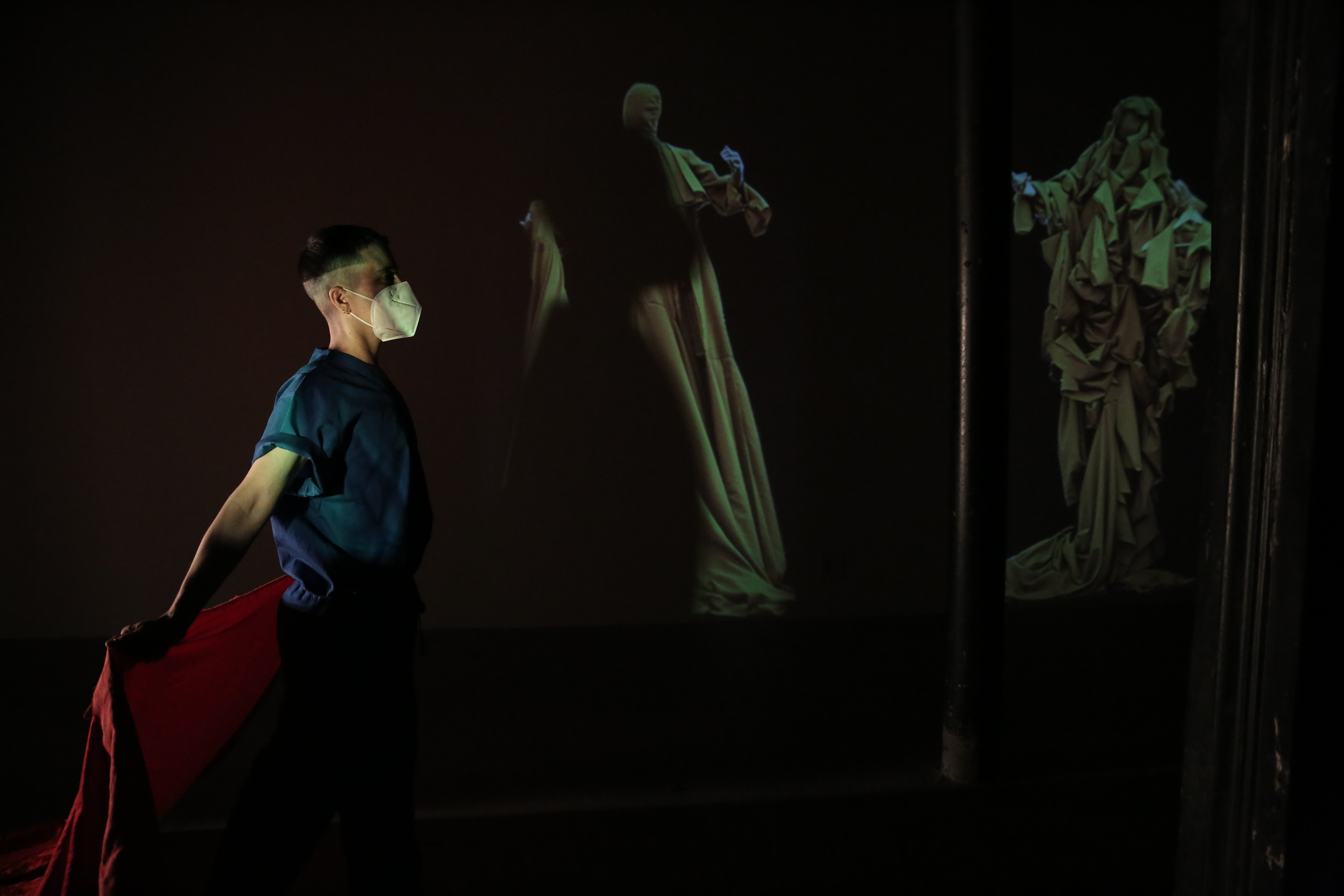 In a darkened space, a person dressed in blue faces to the right holding red fabric behind them. On the back wall are images of figures draped in yellow fabric.