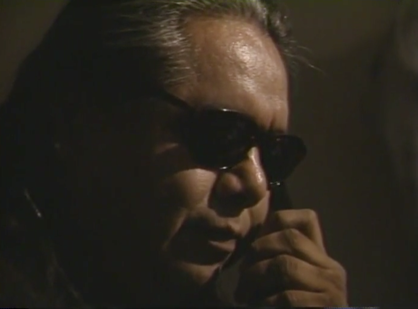 An image of a figure with slightly grey hair who is wearing black sunglasses. They rest their head on their hand while staring downwards.